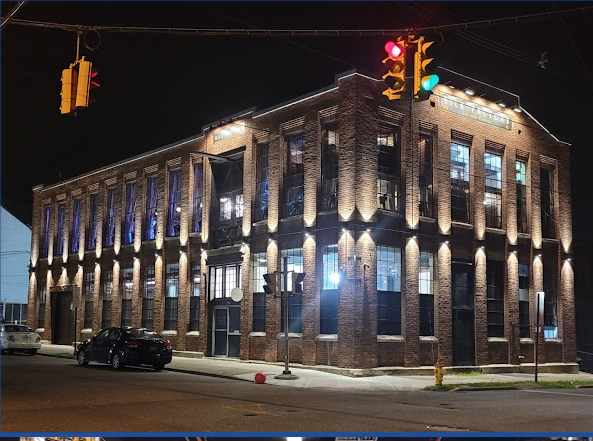 Night view of a The Silk Factory, a well-lit, two-story brick building on a street corner with traffic lights.