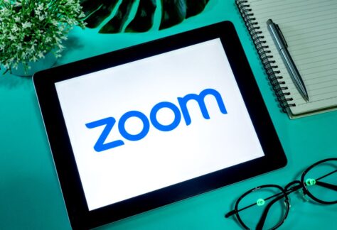Tablet displaying zoom logo on screen, accompanied by glasses, a notepad, and a green plant on a teal surface.