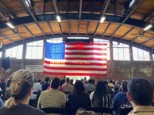 Audience seated in a sports hall, watching a large american flag displayed behind a stage, creating a patriotic backdrop.