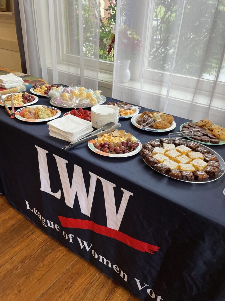 A table with various pastries, fruit, and snacks is set up for an event by the League of Women Voters. Plates, napkins, and utensils are also on the table.