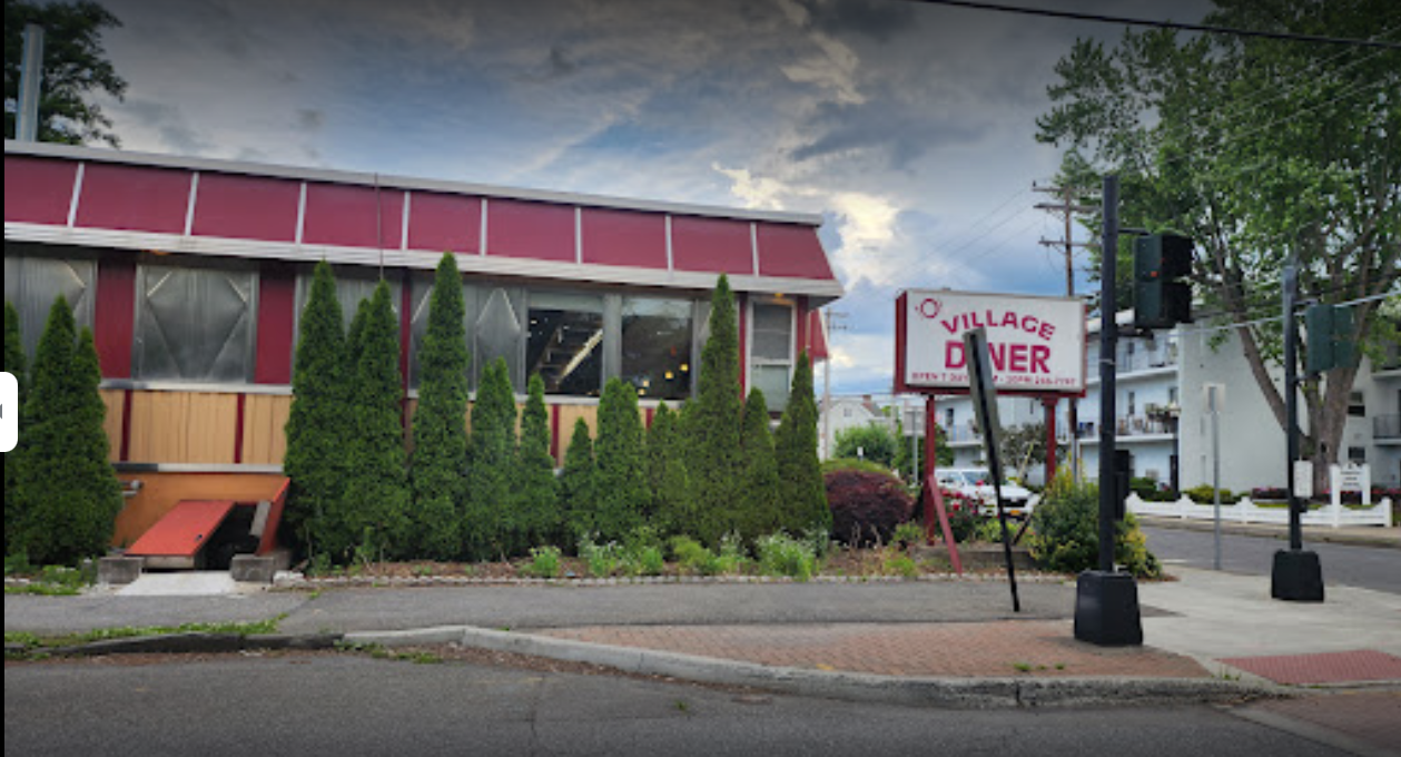 Image of Village Diner with a weathered sign, located on a street corner. The building has a red and silver exterior with tall bushes along the front.