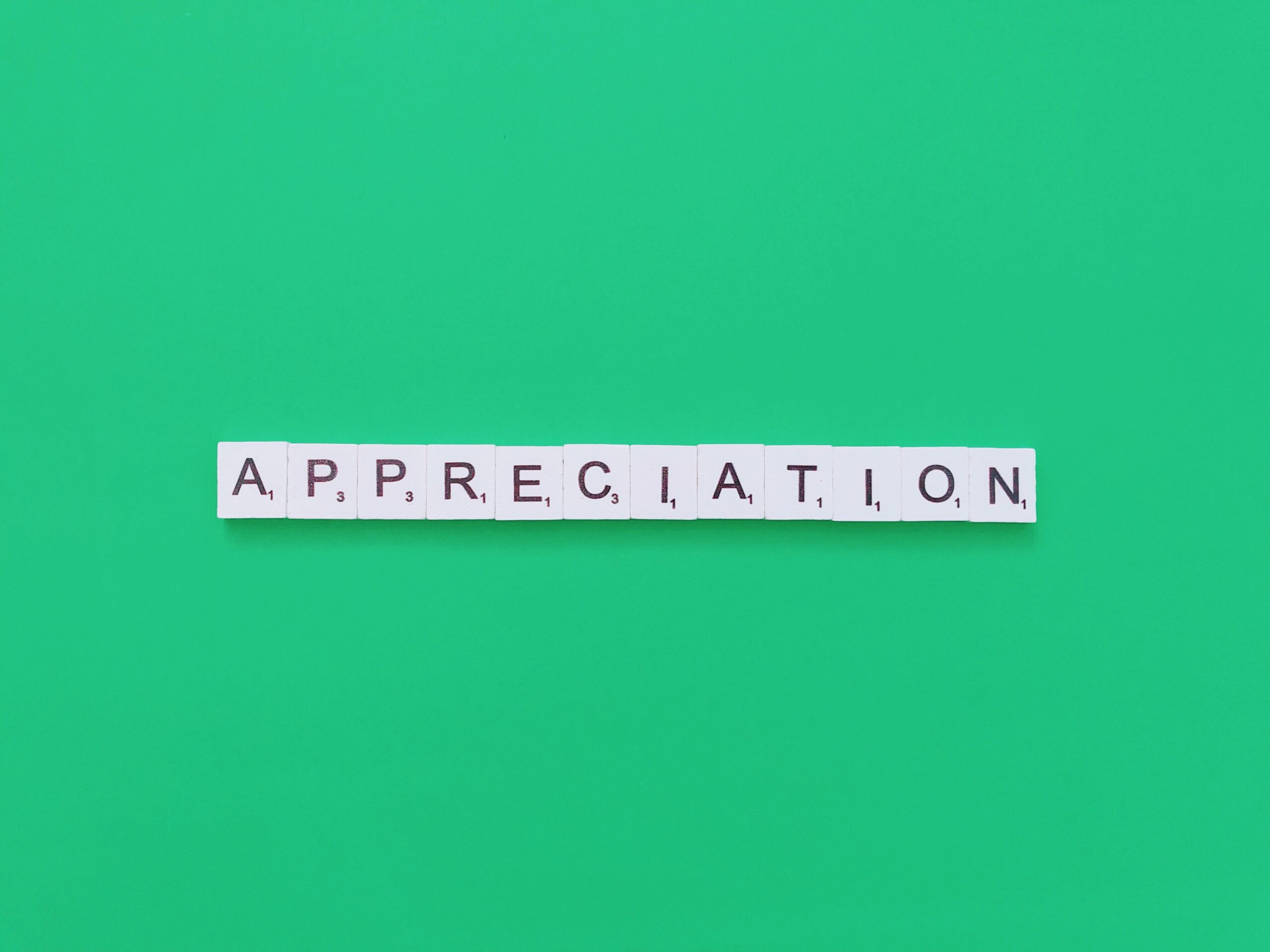 White Scrabble tiles spelling "APPRECIATION" are arranged in a horizontal line against a green background.