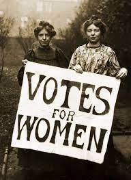 Two women hold a large sign that reads "VOTES FOR WOMEN" during a suffrage movement protest.