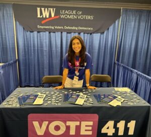 A woman stands behind a table with brochures, buttons, and a "Vote 411" banner at a League of Women Voters booth.