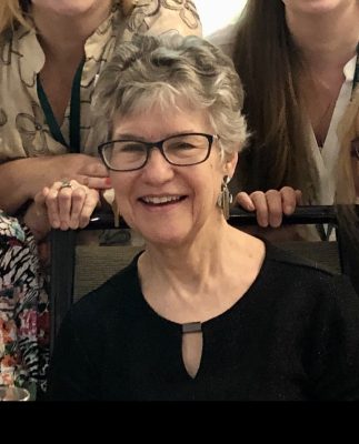 A smiling woman with short gray hair and glasses wearing a black top with a keyhole neckline.