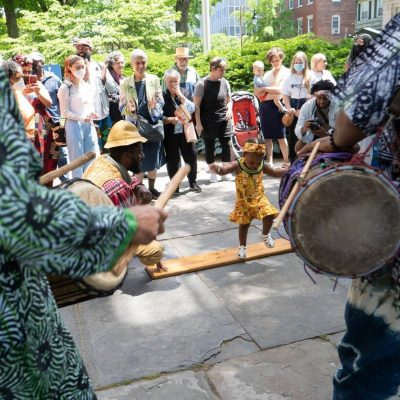 A young child in colorful traditional clothing dances energetically on a wooden plank while surrounded by a group of people, including drummers playing in an outdoor setting.