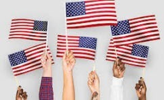 Multiple hands of diverse skin tones raised, each holding a small American flag against a plain background.